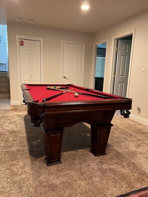 A pool table with a red felt surface, surrounded by carpeted flooring, in a room with white doors and a visible window.