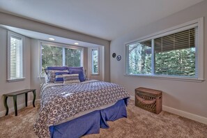Une chambre avec un grand lit, deux fenêtres équipées de stores, un petit banc et un panier en osier.