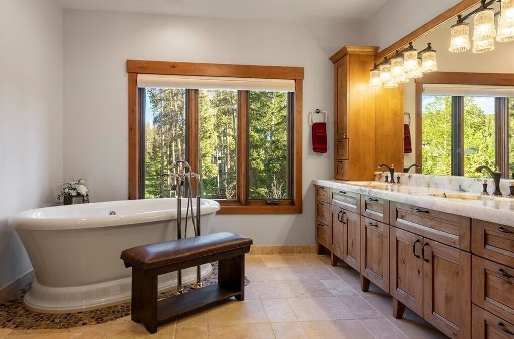 A bathroom with a freestanding bathtub, wooden cabinets, a bench, and a large window with a view of trees.