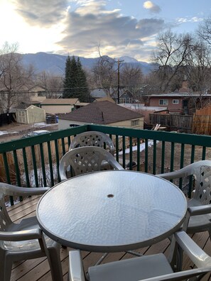 A deck with a round table and chairs, overlooking a snowy landscape with houses and mountains in the background.