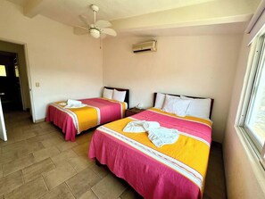 A hotel room with two beds, a ceiling fan, an air conditioning unit, and a window.