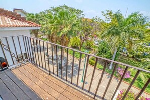 A balcony with a wooden floor and metal railing overlooking a garden with palm trees and other greenery.