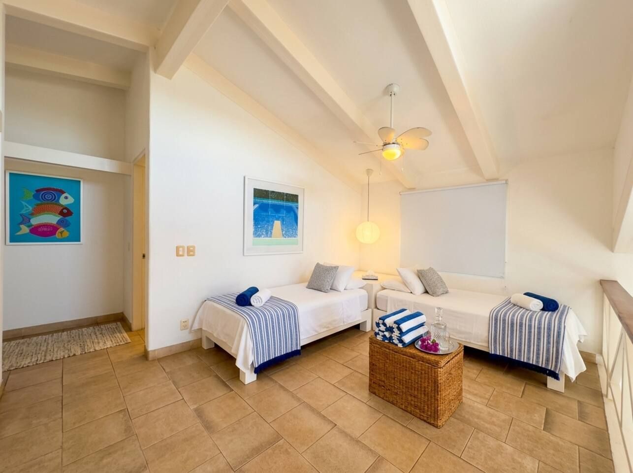 Ein Zimmer mit zwei Betten, einem Deckenventilator, einer Wandleuchte und einem kleinen Tisch mit einem Korb.