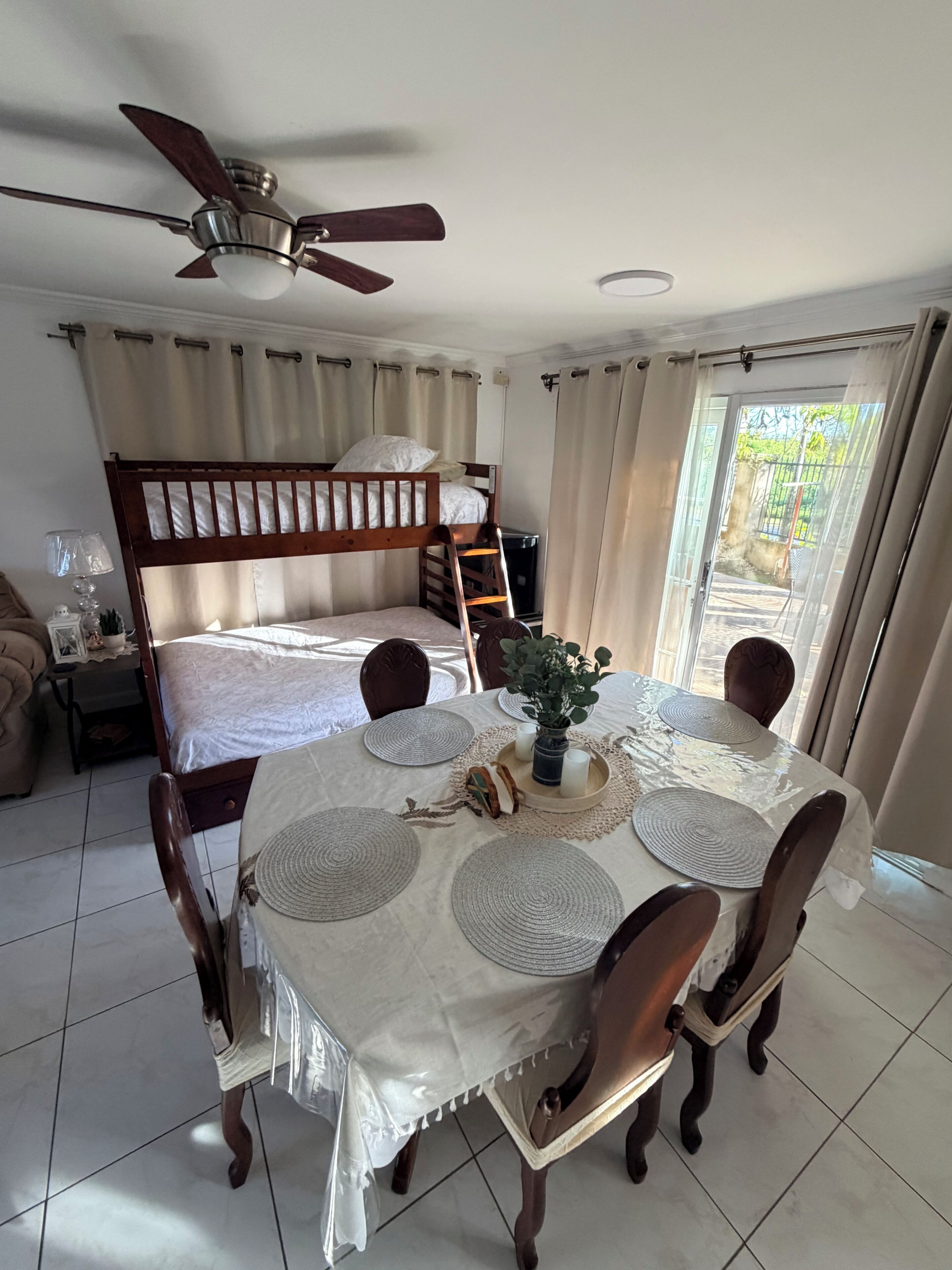 A room with a bunk bed, a dining table with placemats, chairs, a ceiling fan, and a view through a sliding glass door.
