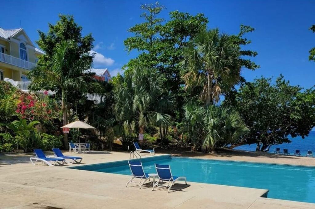 A pool area with lounge chairs, a clear view of the ocean, and lush greenery.