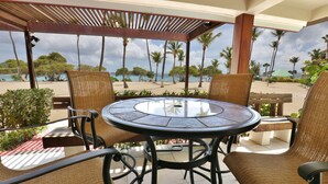 Ein überdachter Außenbereich mit Korbstühlen und einem Tisch mit Glasplatte, mit Blick auf einen Strand mit Palmen und einem klaren Himmel.