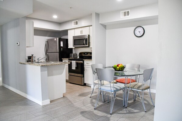 A modern kitchen with a built-in refrigerator, microwave, and oven. There is a dining area with a table and chairs, and a clock on the wall.