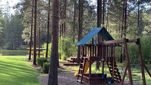 Ein Spielplatz mit einer Holzschaukel und Rutschen, umgeben von Bäumen und einer Rasenfläche.