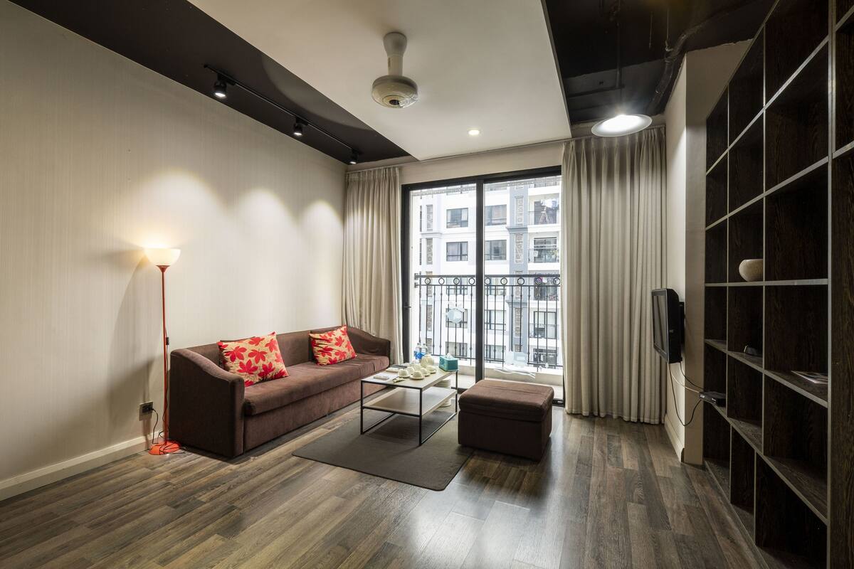 A modern living room with a sofa, coffee table, and ottoman, a bookshelf, and a large window with curtains.