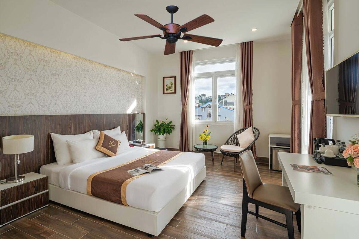 Une chambre d’hôtel avec un grand lit, un bureau, une chaise, un ventilateur de plafond et une fenêtre avec des rideaux.
