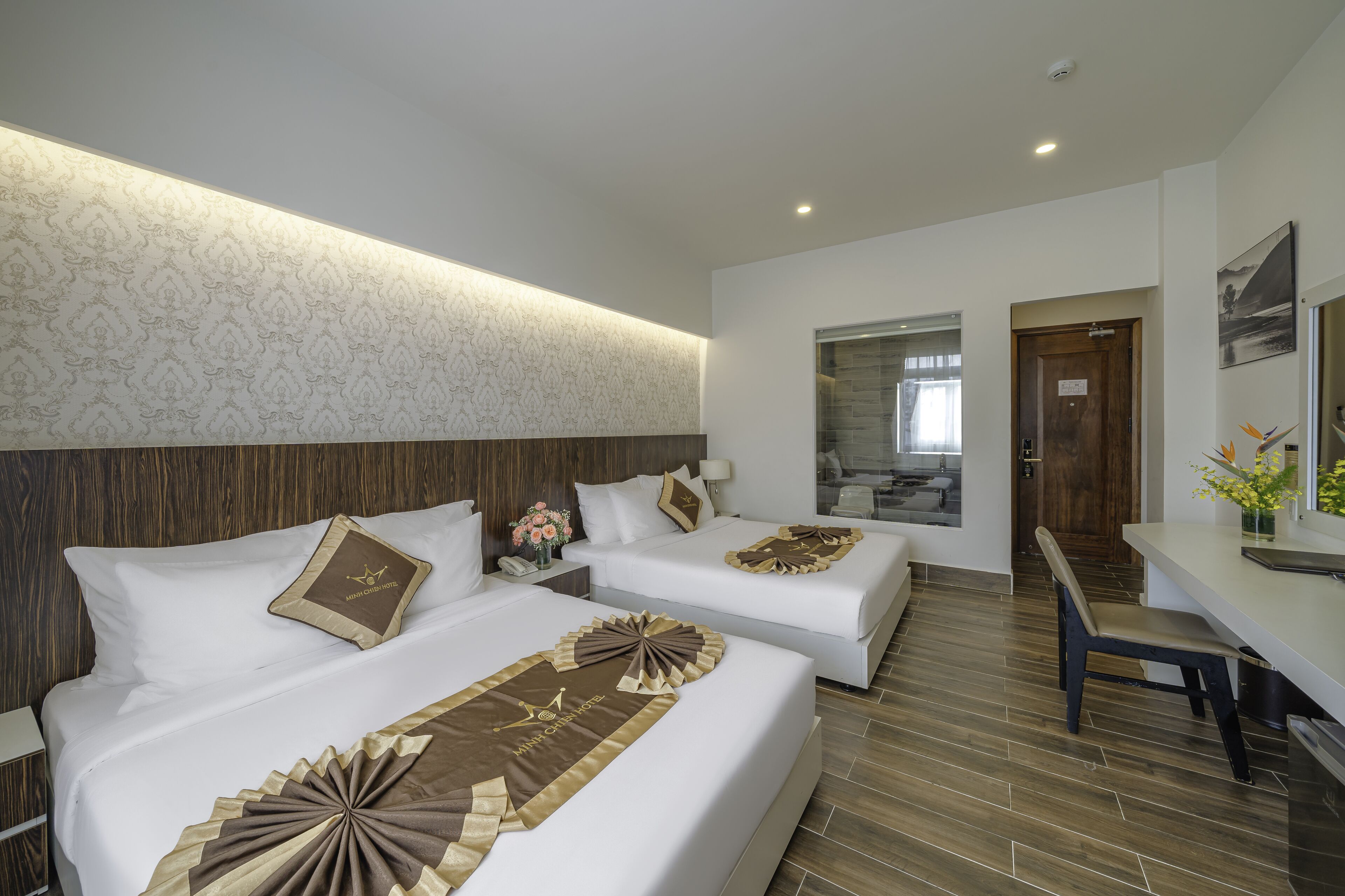 A hotel room with two beds, a desk, and a bathroom visible through a glass door.