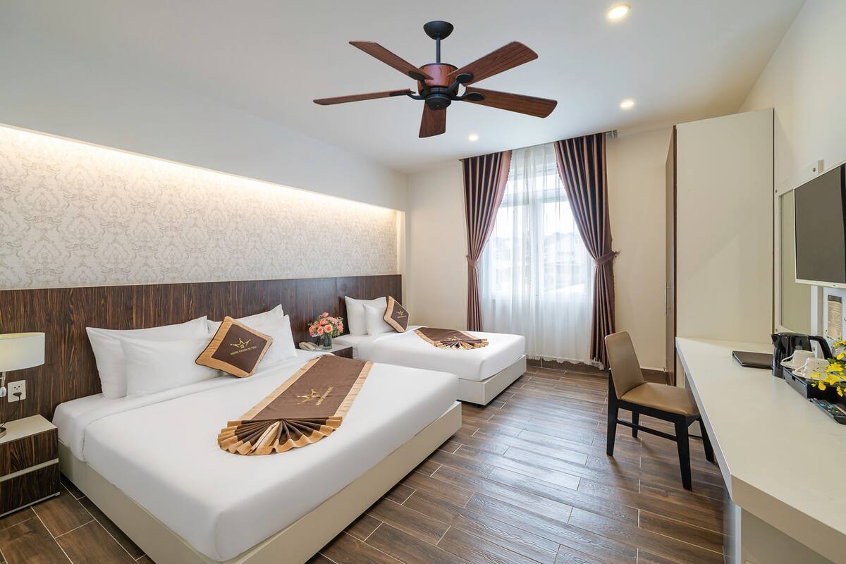 Une chambre d’hôtel avec deux lits, un bureau, une chaise, un ventilateur de plafond et une grande fenêtre avec des rideaux.