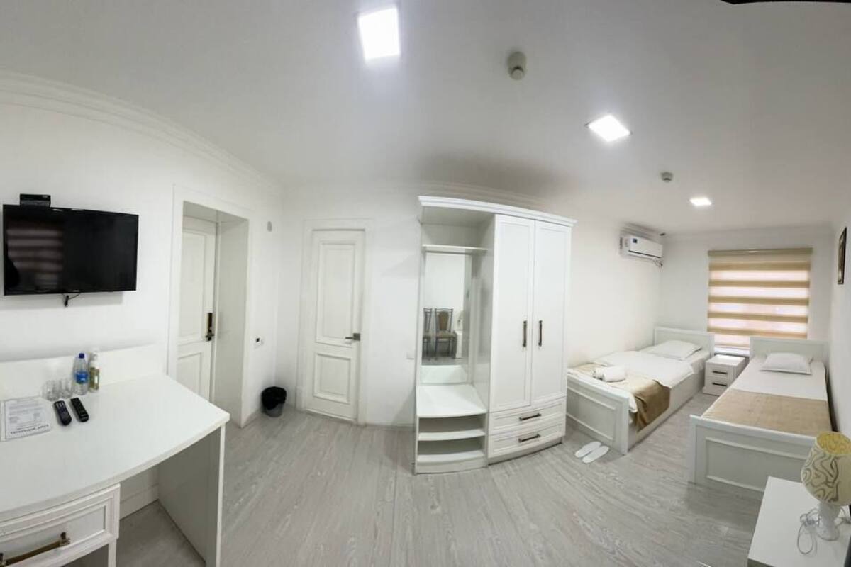 A modern bedroom with a white wardrobe, a bed with a beige headboard, a bedside table, and a flat-screen TV mounted on the wall.