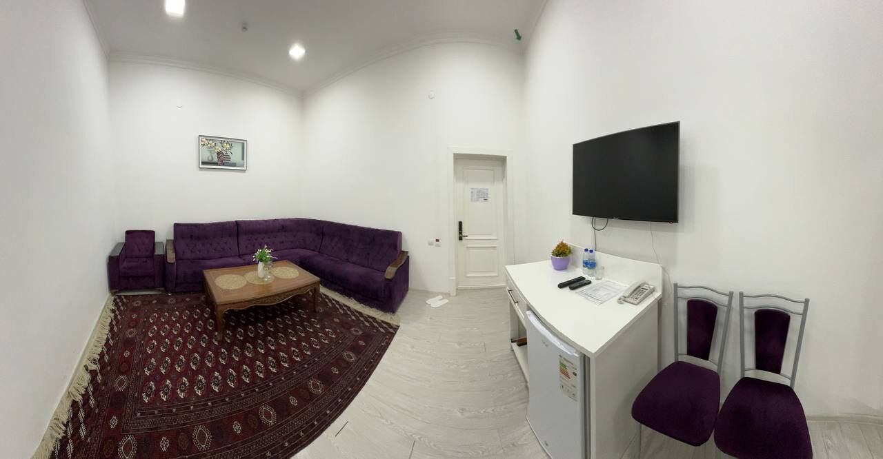 A living room with a purple sofa set, a coffee table, a flat-screen TV mounted on the wall, and a small desk with two chairs.