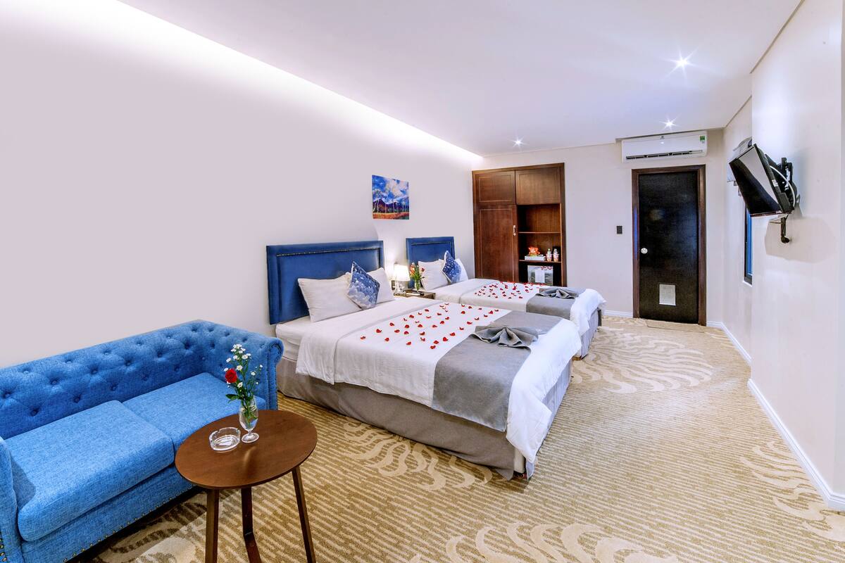 A hotel room with two beds, a blue sofa, a small table with a vase of flowers, and a carpeted floor.