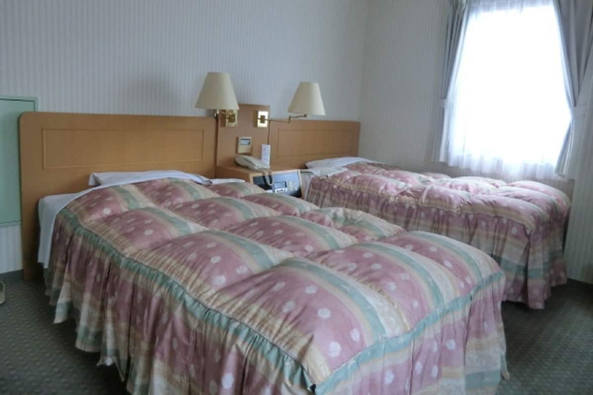 Quarto de hotel com duas camas, cada uma com colcha estampada com flores, cabeceira de madeira, duas luminárias de cabeceira, mesa de cabeceira com telefone e uma janela com cortinas.