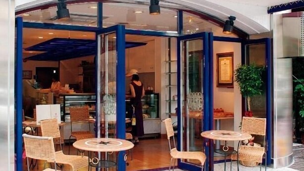 Un restaurante con toldo azul y blanco a rayas, puertas de cristal y terraza con sillas y mesas de mimbre.
