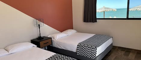 A hotel room with two beds, a nightstand, a window with a view of the sea, and a brown wall.