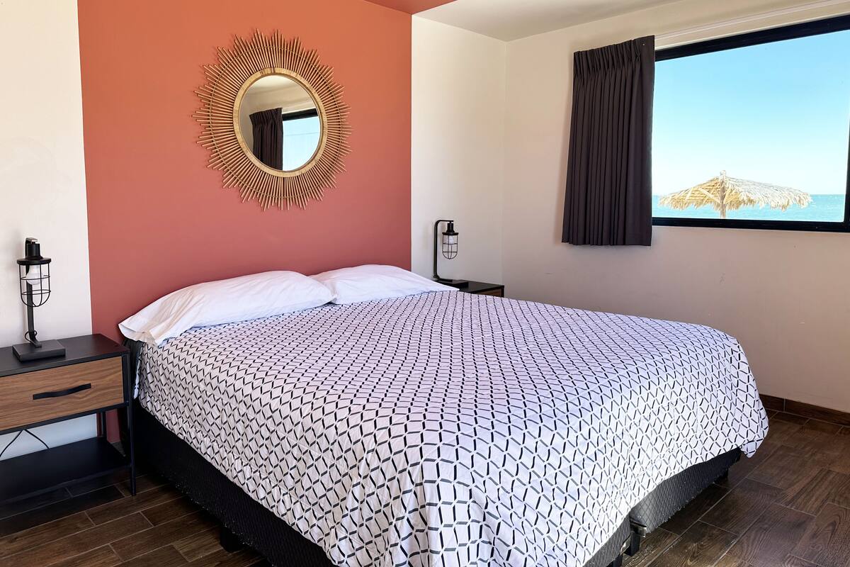 A bedroom with a bed, a nightstand, a lamp, a mirror, and a window with a view of the sea.