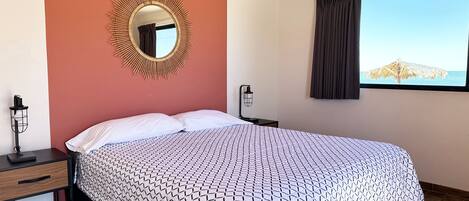 A bedroom with a bed, a nightstand, a lamp, a mirror, and a window with a view of the sea.