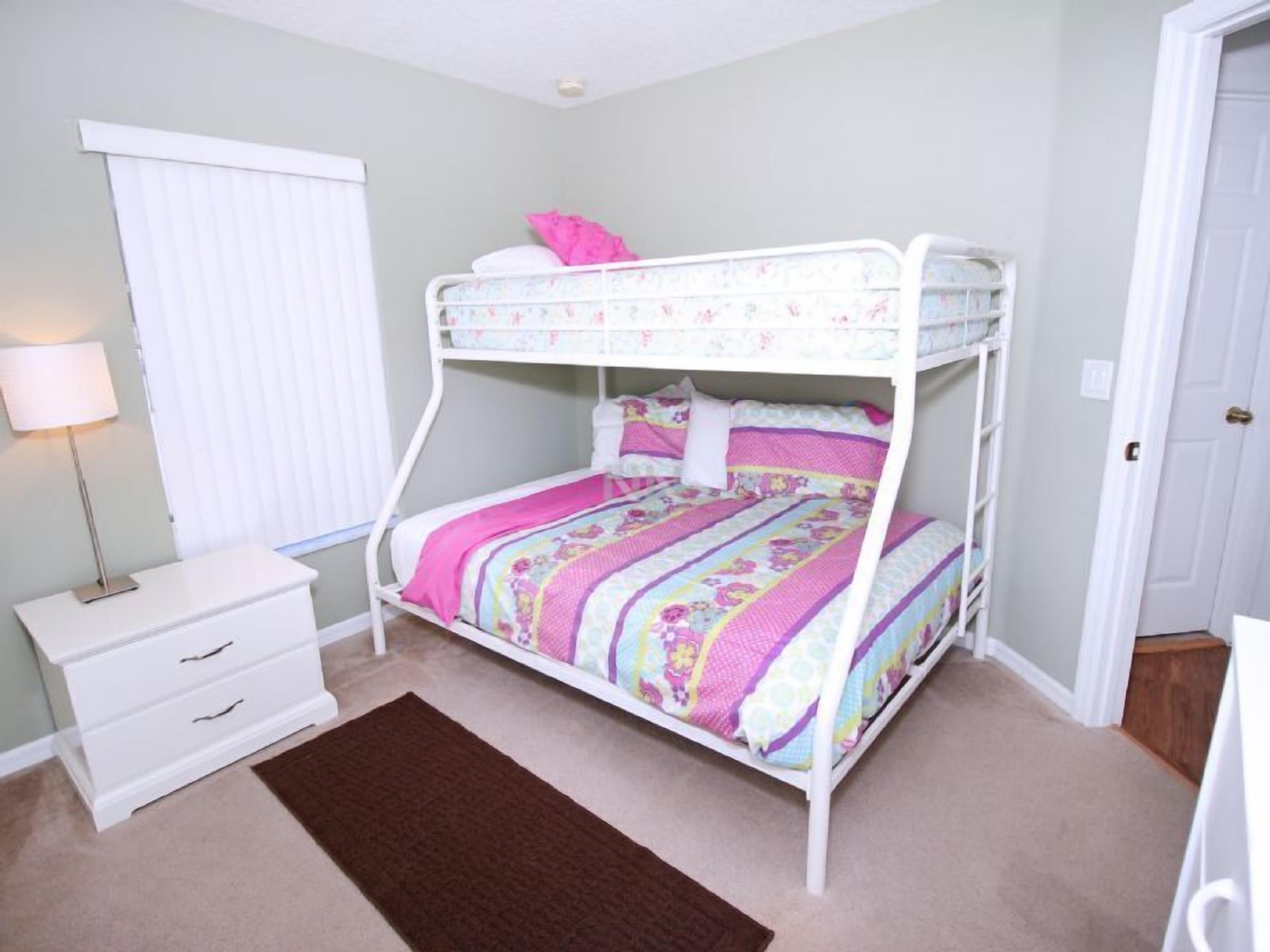 A bunk bed with a floral bedspread, a bedside table with drawers, a lamp, and a window with blinds.
