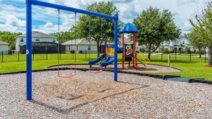 Ein Spielplatz mit Schaukeln und Rutschen, umzäunt und mit Häusern im Hintergrund.
