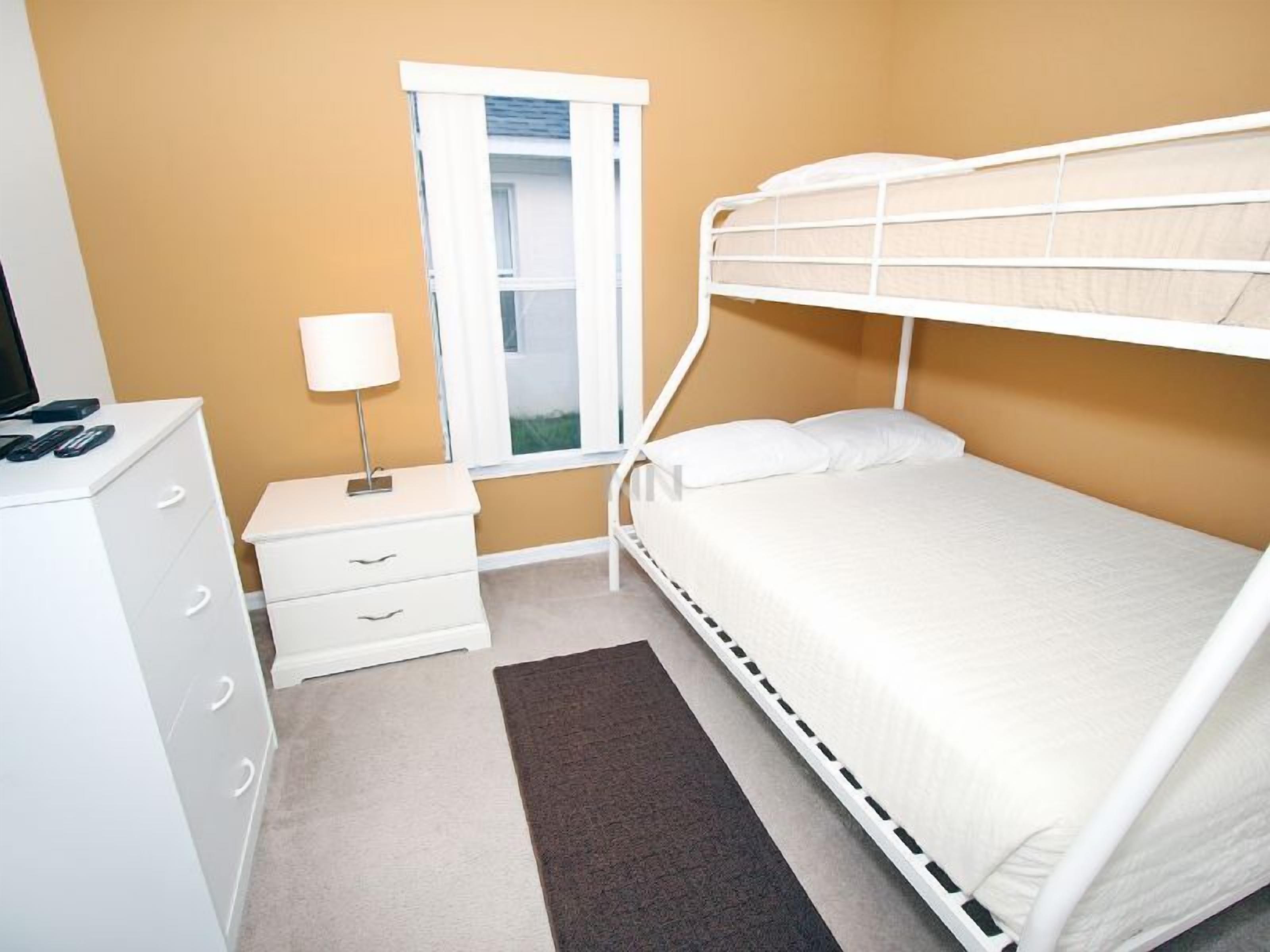 Une chambre avec un lit superposé, une table de chevet avec une lampe, une commode et une fenêtre avec des rideaux.