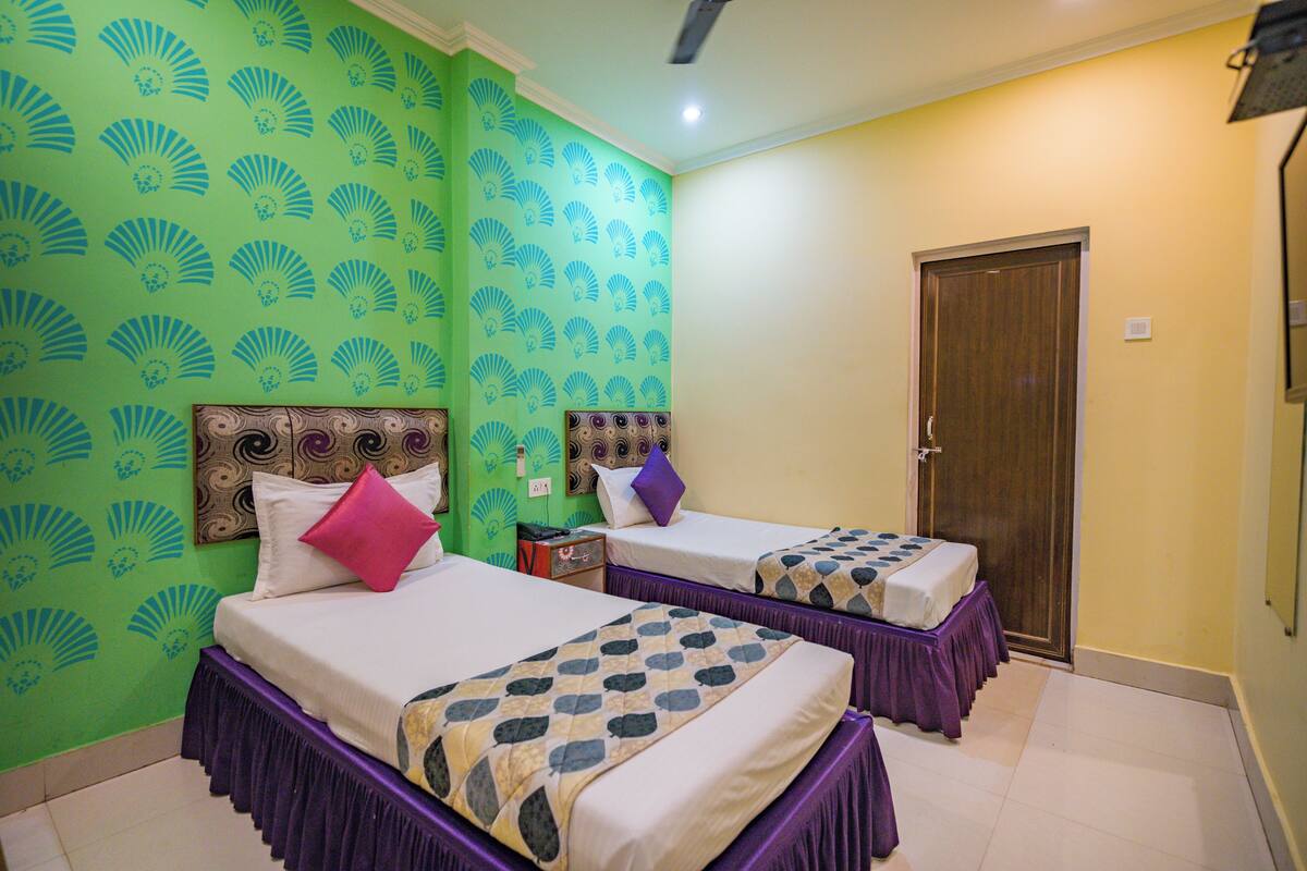 A hotel room with two beds, each with a purple skirted base and a patterned bedspread. The walls are green with a fan pattern, and there is a wooden door.