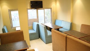 A room with a TV mounted on the wall, a table, and two seating areas with cushions. - Hotel Samman (Varanasi)