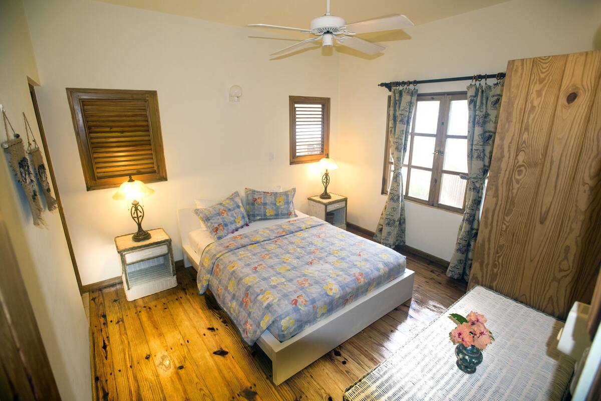 A bedroom with a bed, two bedside tables, a ceiling fan, a window with curtains, and a wooden floor.