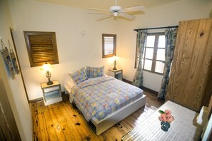 Un dormitorio con una cama, dos mesitas de noche, un ventilador de techo, una ventana con cortinas y piso de madera.