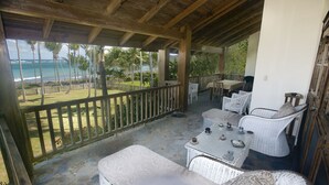 A covered outdoor seating area with wicker furniture, a stone floor, and a view of the ocean and palm trees. - Villa Natasha (Las Galeras)