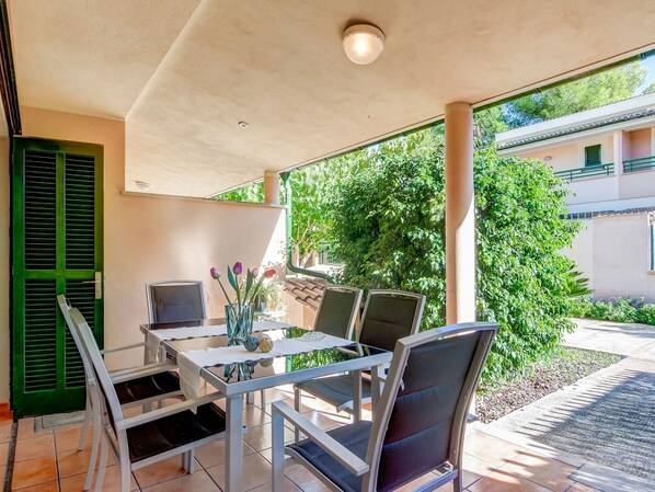 Eine überdachte Terrasse mit Tisch und Stühlen, eine grüne Tür sowie Blick auf ein Gebäude und Bäume.