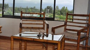 Two bamboo chairs with woven seats and backs, a glass table with a remote control, and a view of greenery and mountains through large windows. - Peaks Hotel Limited (Nanyuki)