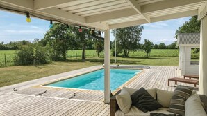 Eine Holzterrasse mit Pool, einem überdachten Sitzbereich und Blick auf eine grüne Landschaft.