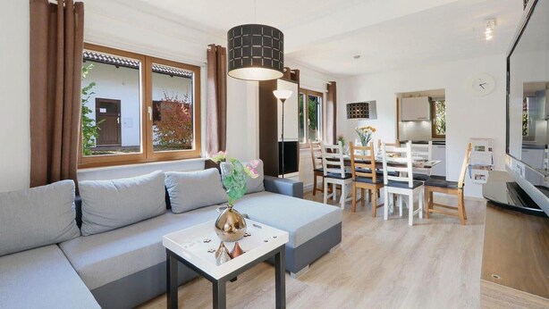 A modern living room with a sofa, a dining table with chairs, a kitchen area, and large windows with curtains.