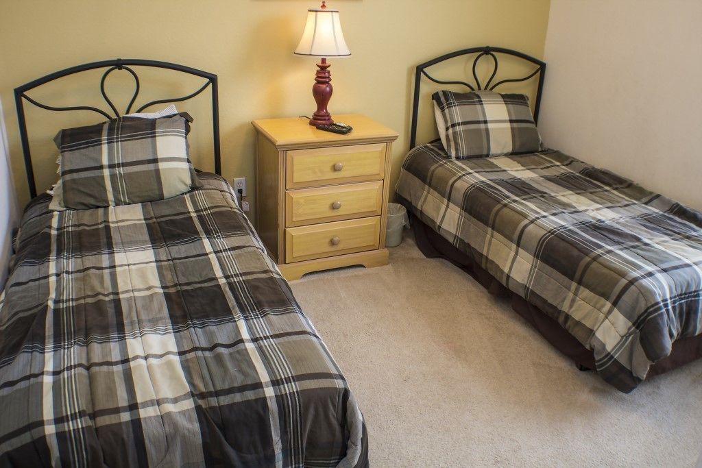 A twin bedroom with two beds, each with a plaid duvet, a bedside table with a lamp, and a small drawer.