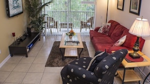 A living room with a red sofa, a black armchair, a glass coffee table, and a floor lamp. There is a large window with a view of greenery outside. - Ip60127 - Windsor Palms Resort - 3 Bed 2 Baths Condo (Kissimmee)