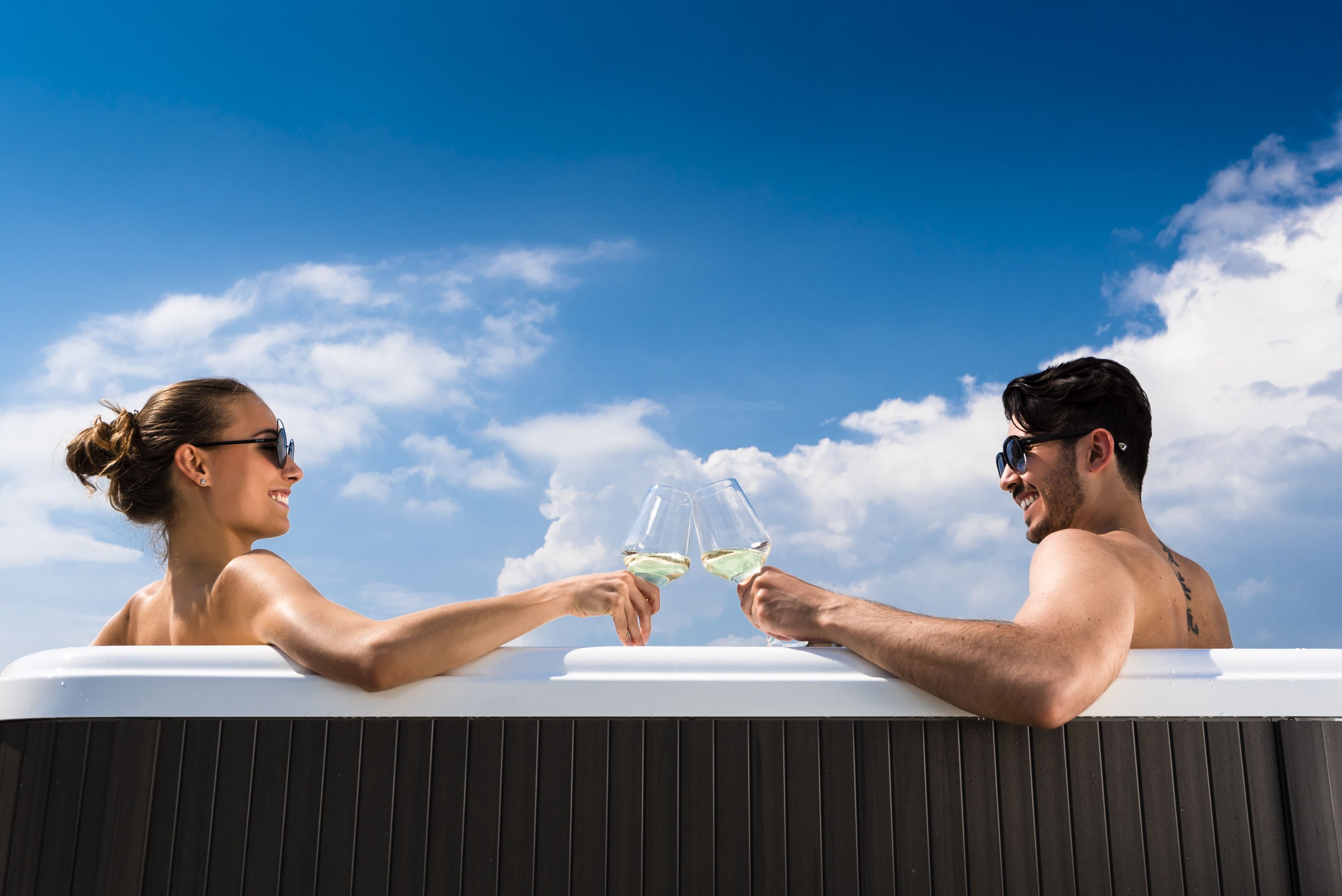 Two people toasting with wine glasses in a hot tub against a clear blue sky.