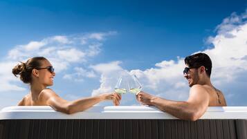 Two people toasting with wine glasses in a hot tub against a clear blue sky.