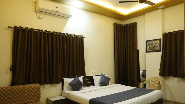 A hotel room with a bed, a chair, a small table, and an air conditioning unit. - Balaji Lodging (Pandharpur)