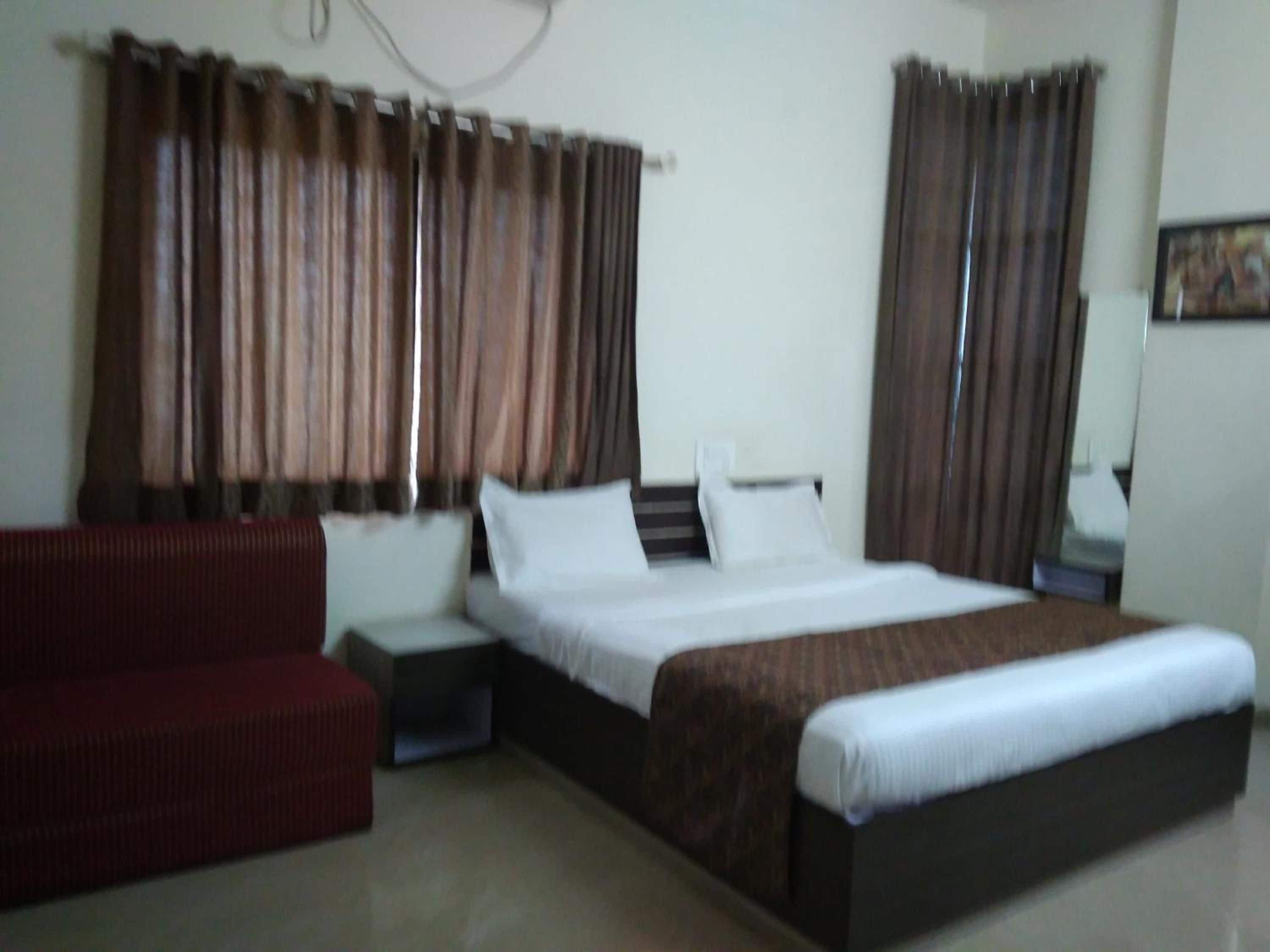 A hotel room with a bed, a red sofa, a nightstand, and curtains.