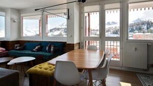 En moderne stue med et træspisebord, hvide stole, en blå sofa og en gul fodskammel. Store vinduer giver udsigt til et sneklædt bjerglandskab.