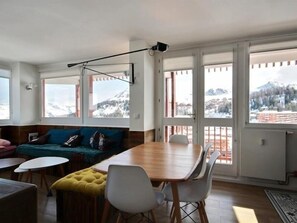 Un soggiorno moderno con un tavolo da pranzo in legno, sedie bianche, un divano blu e un poggiapiedi giallo. Ampie finestre offrono vista su un paesaggio montano innevato.