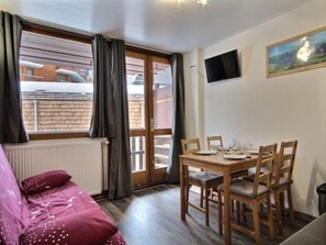 Ein gemütliches Zimmer mit einem Holzesstisch, gedeckt für vier Personen, einer Couch mit gemustertem Bezug, einem Fernseher an der Wand und einem Fenster mit Vorhängen.
