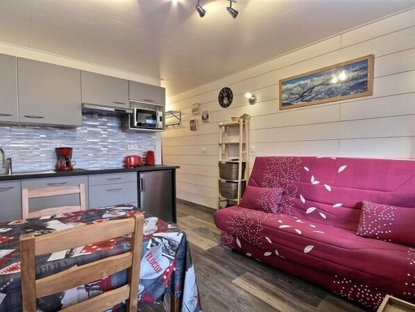 Ett kompakt bostadsutrymme med ett litet kök, en röd soffa, ett matbord med ett mönstrat bordsduk och en väggklocka.
