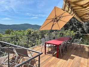 Una terraza de madera con una mesa y sillas rojas, un paraguas color crema y vistas a colinas verdes.