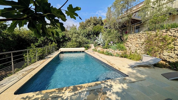 A rectangular swimming pool with a stone wall and a metal railing on one side, surrounded by greenery and a stone building.