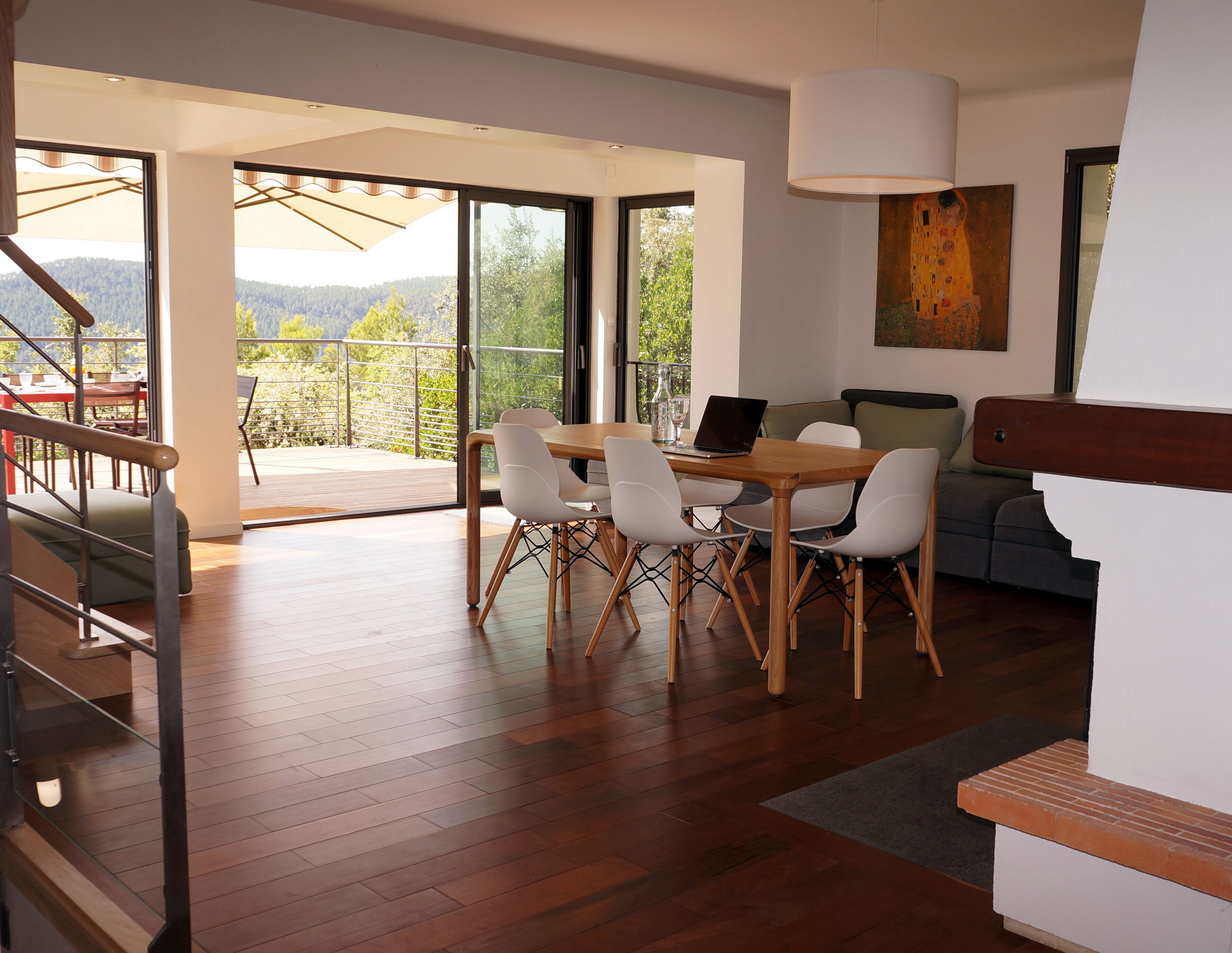 Un salon moderne avec une table à manger en bois, des chaises blanches et un canapé. Il y a un balcon avec vue sur l’extérieur.