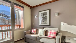 Ein gemütliches Wohnzimmer mit einer Couch, Kissen, einem gerahmten Bild und Blick auf einen verschneiten Berg durch eine Schiebetür.